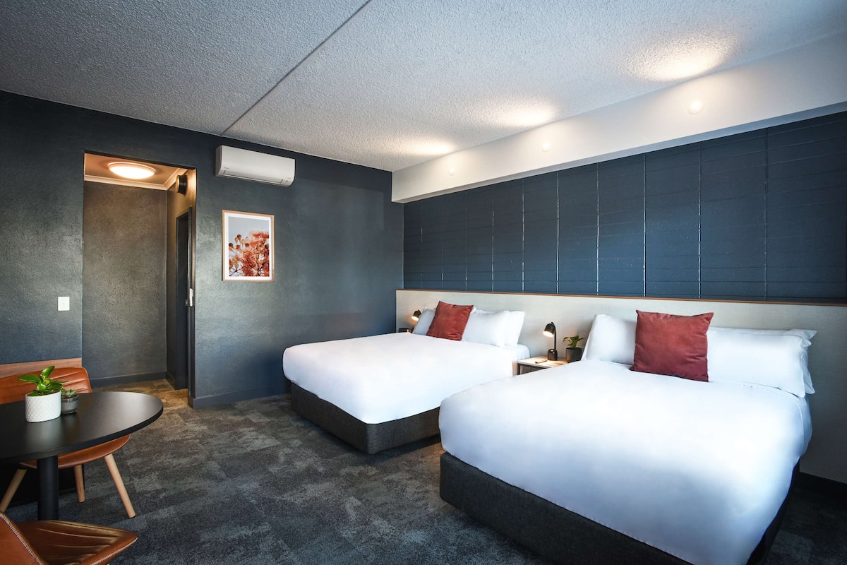 Une chambre d’hôtel avec deux lits, une petite table avec des chaises, une plante en pot et un tableau encadré au mur.