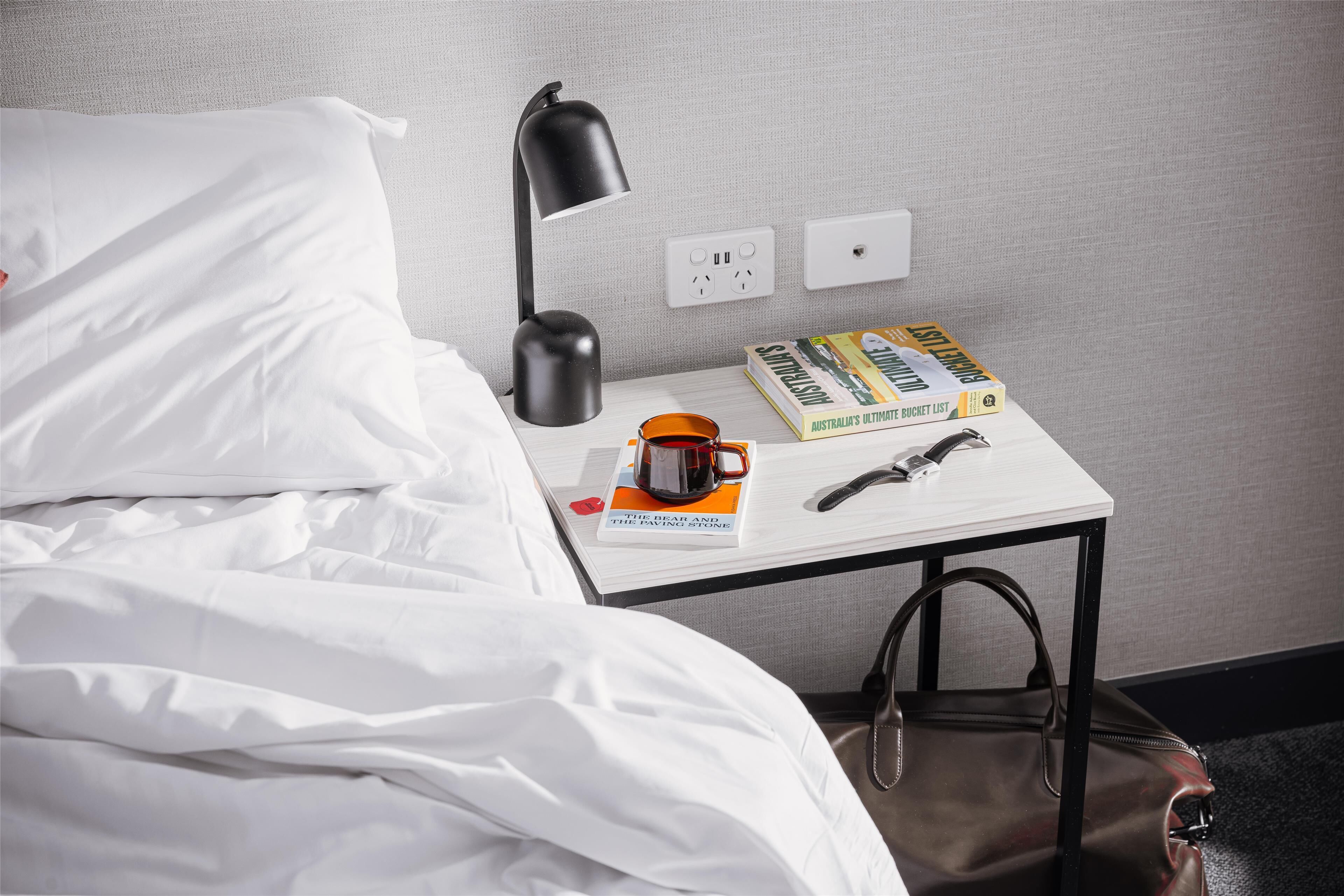 A neatly made bed with white linens, a black desk lamp, a book titled "Australia's Ultimate Bucket List," a mug, and a pair of glasses.