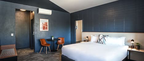 Une chambre d’hôtel moderne avec un grand lit, un bureau avec deux chaises, une petite table avec une plante et un climatiseur fixé au mur.