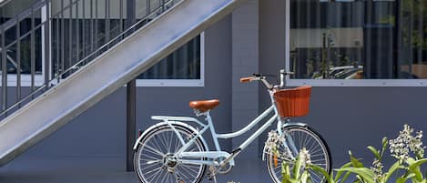 A white bicycle with a brown seat and orange basket, parked in front of a building with a grey facade and a metal staircase.