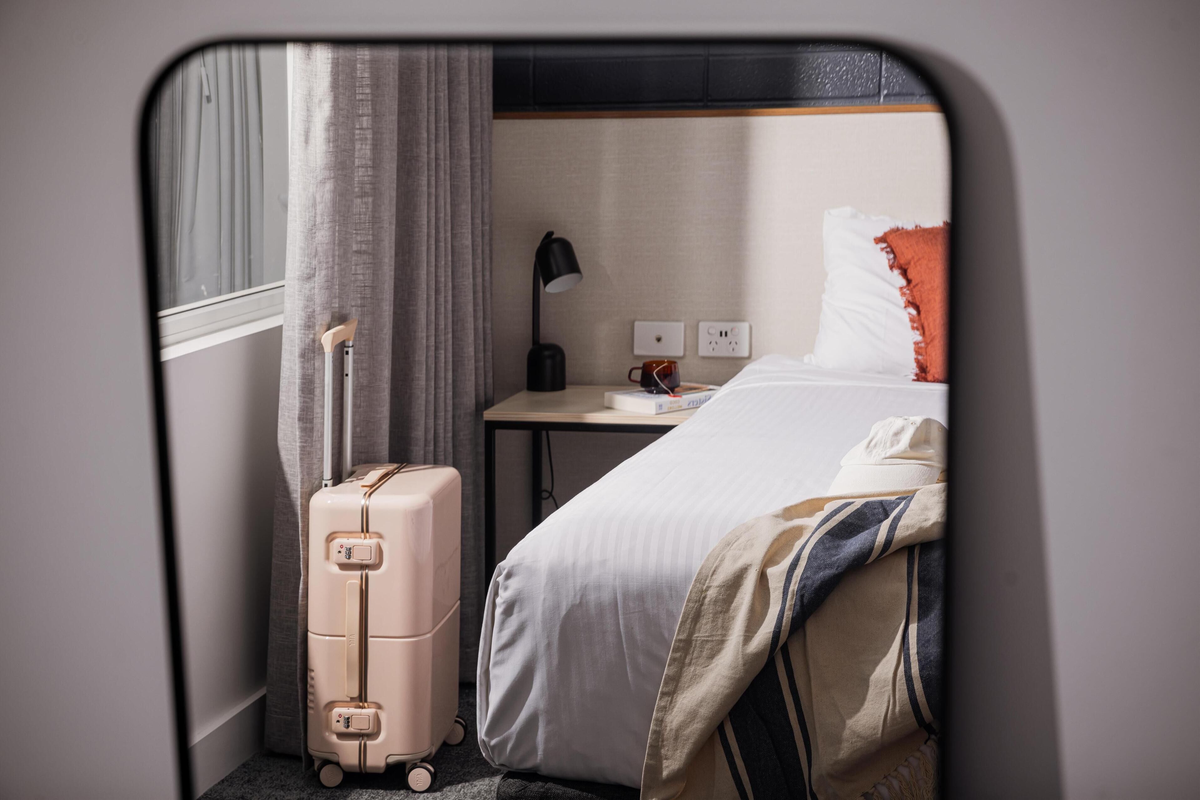 A hotel room with a bed, a bedside table with a lamp, a suitcase, and a window with curtains.