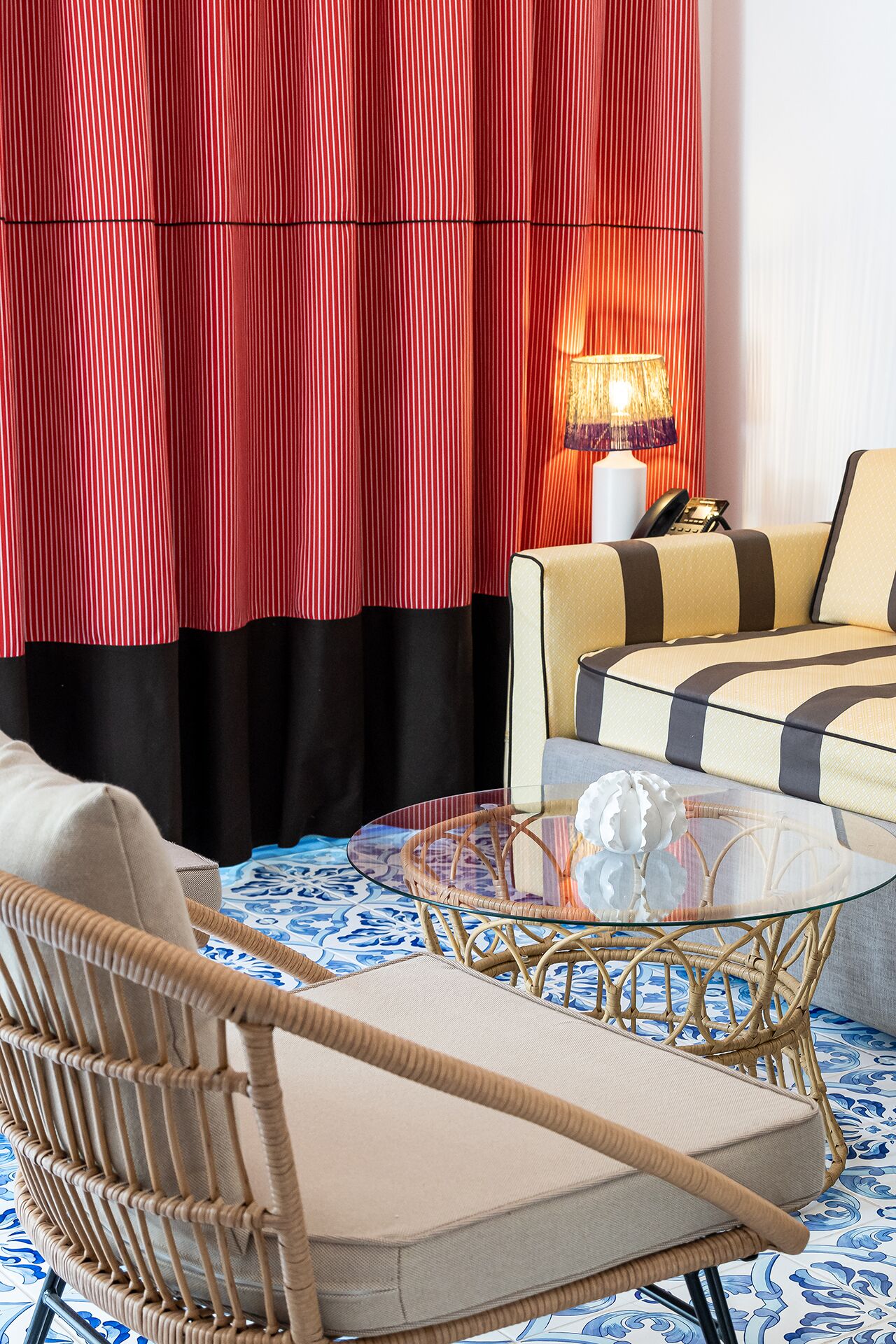A room with a wicker chair, a sofa with striped cushions, a glass coffee table, and a lamp.