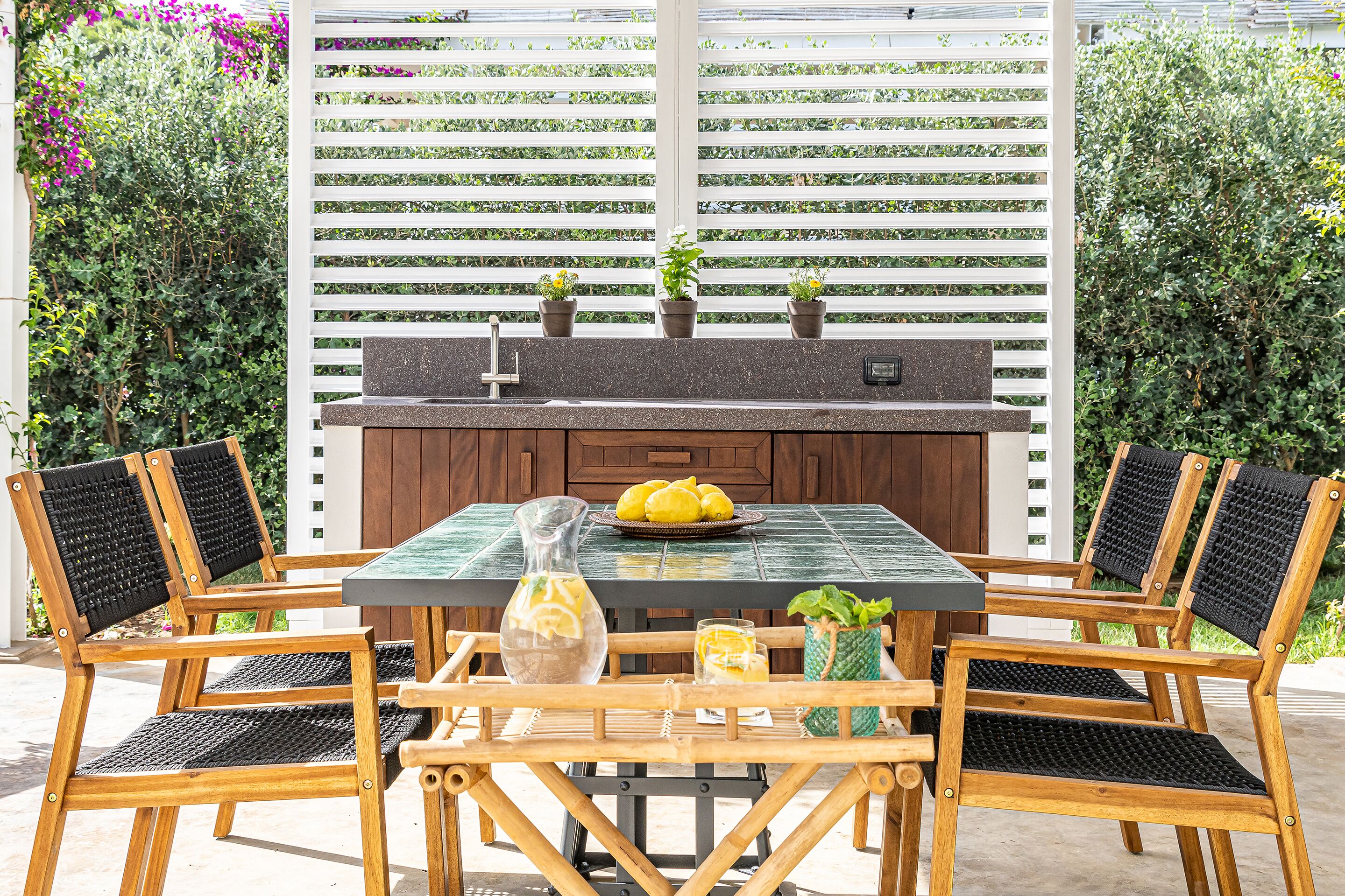En udendørs terrasse med et træbord og stole, grill og krukker med planter.