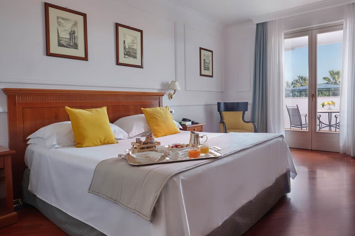 Hotelkamer met een groot bed, twee gele kussens, een dienblad met ontbijtartikelen en een balkon met buitenmeubilair.