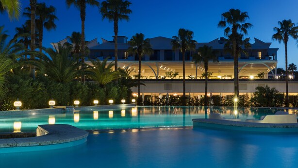 A large outdoor swimming pool with multiple sections, surrounded by palm trees and a well-lit building in the background. - Savoy Hotel & Spa (Capaccio-Paestum)