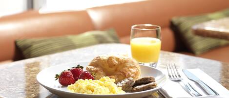 Un desayuno con huevos revueltos, salchicha, un croissant y fresas, un vaso de jugo de naranja, y un tenedor y cuchillo sobre una mesa.