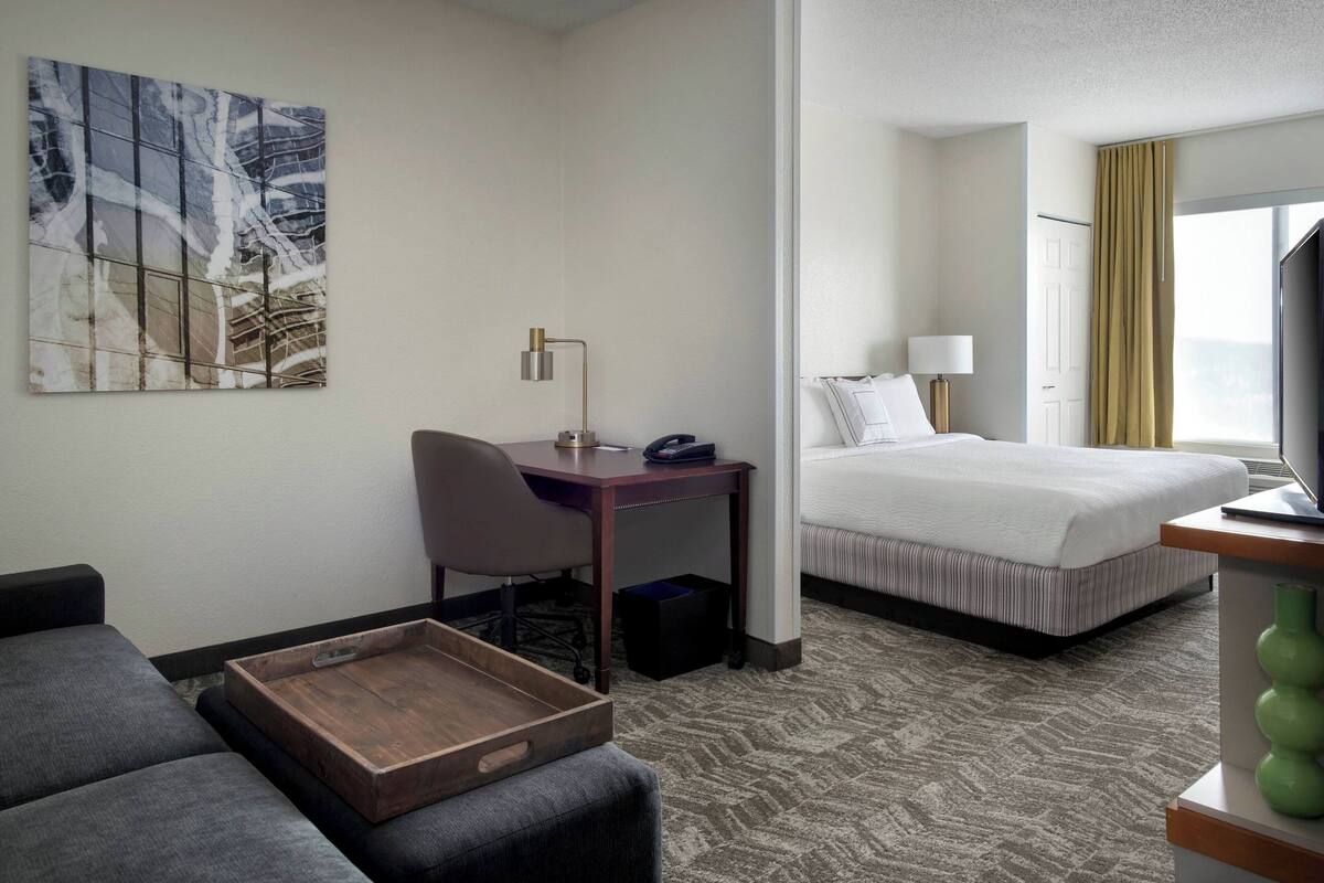 A hotel room with a bed, a desk, a chair, a small table, a painting on the wall, and a window with curtains.