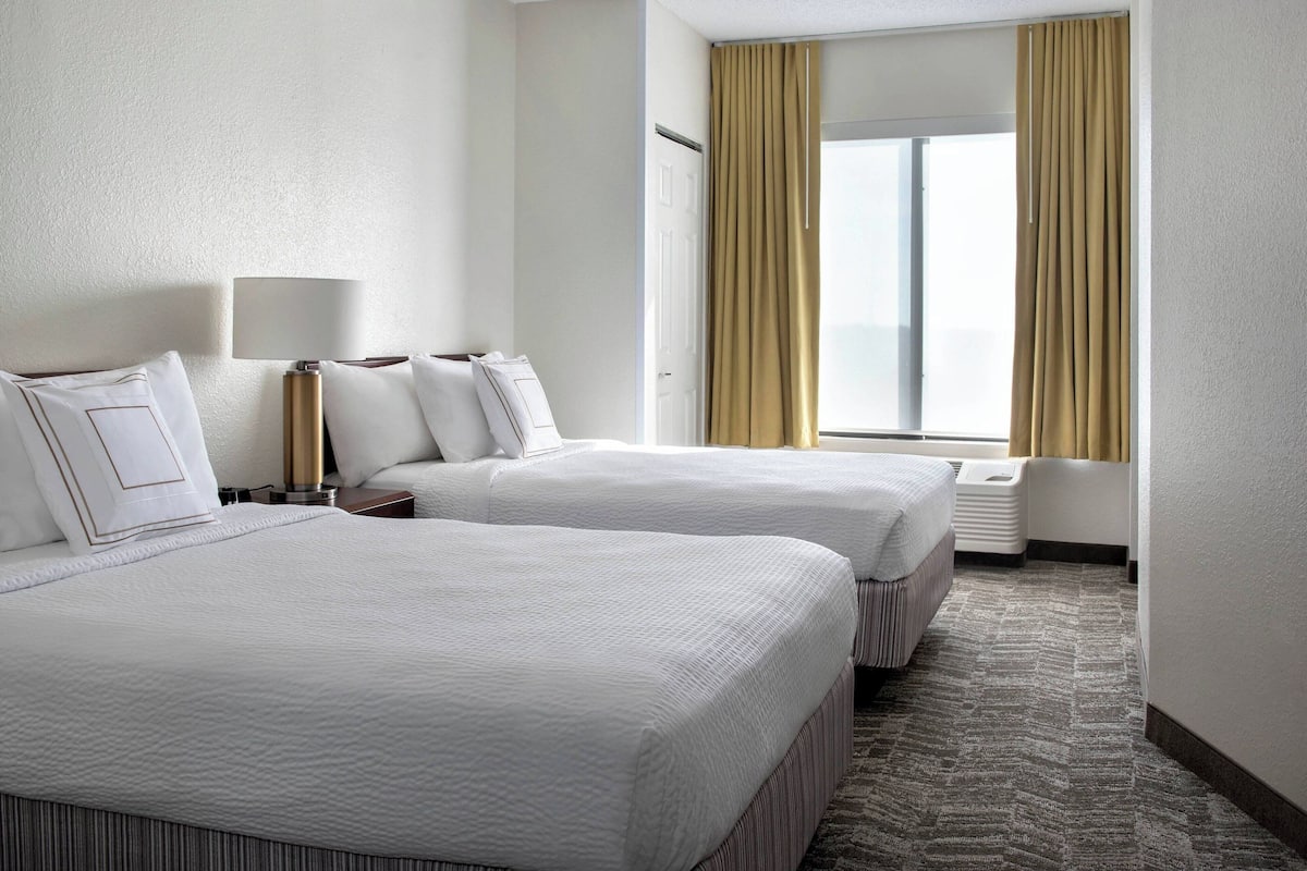 A hotel room with two beds, a bedside table with a lamp, a window with curtains, and a carpeted floor.