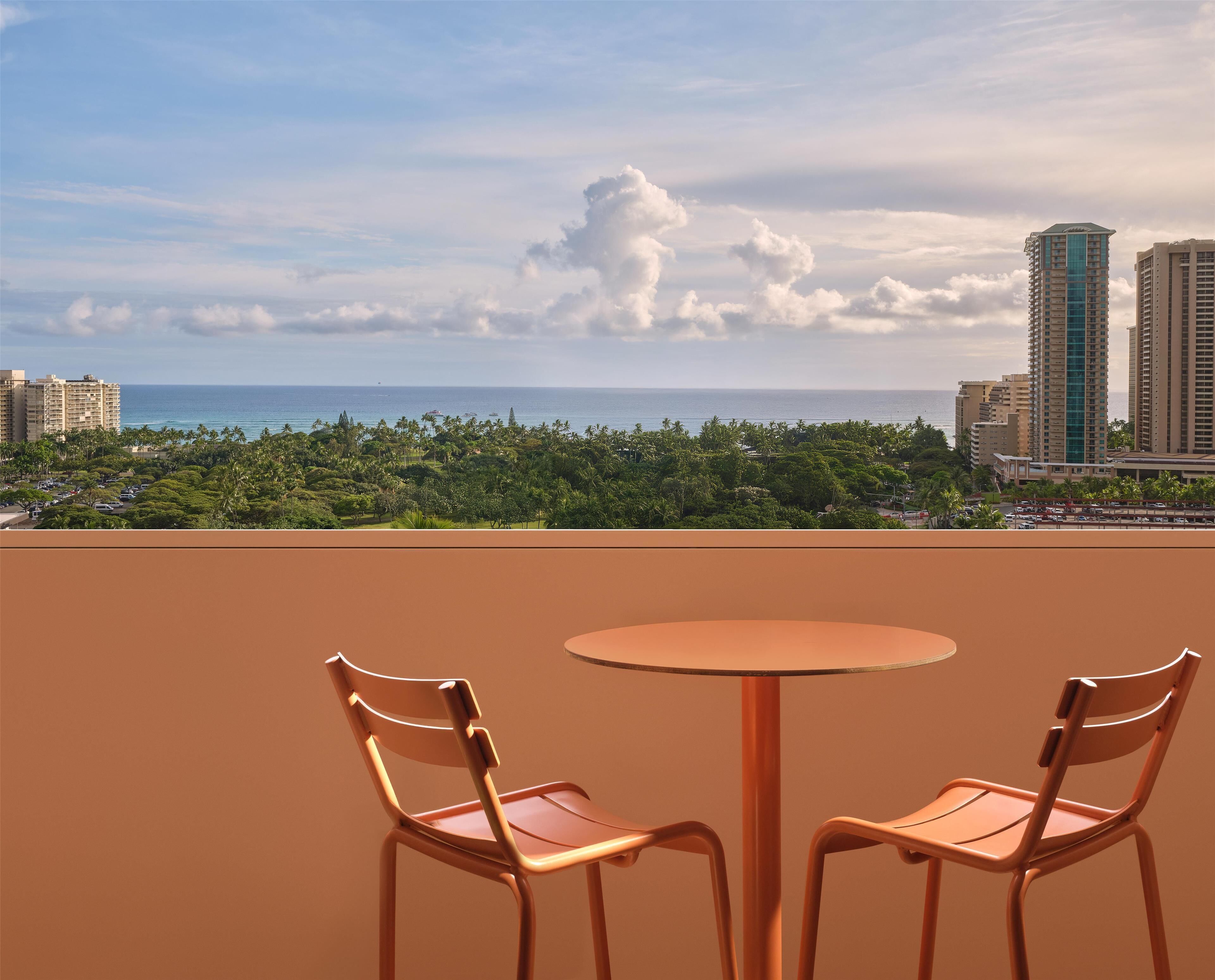 A balcony with two chairs and a table, overlooking a cityscape with tall buildings and a view of the ocean.