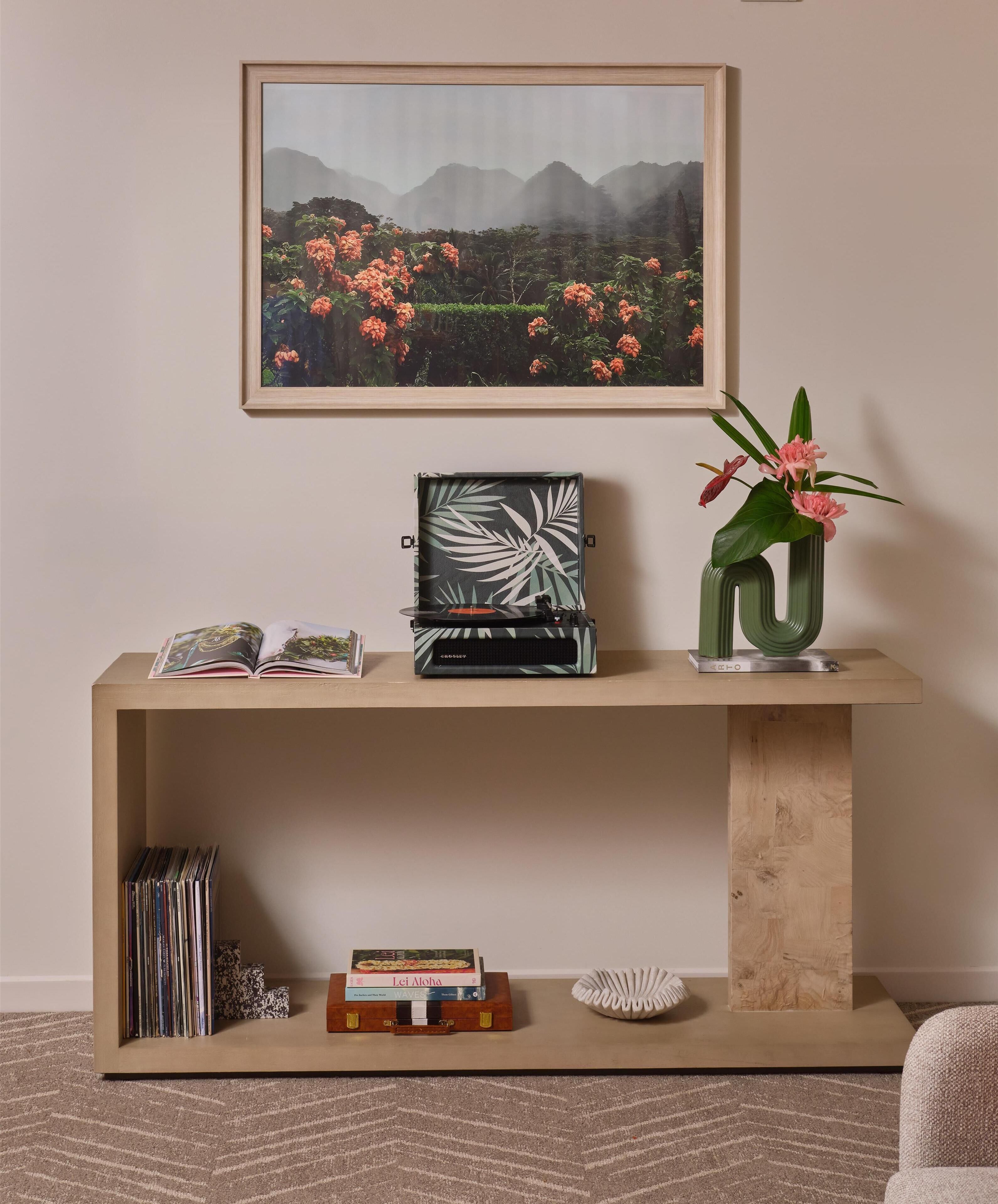 A wooden console table with a framed landscape painting, a vinyl record player, a vase with pink flowers, and a small book titled "Lei Aloha".