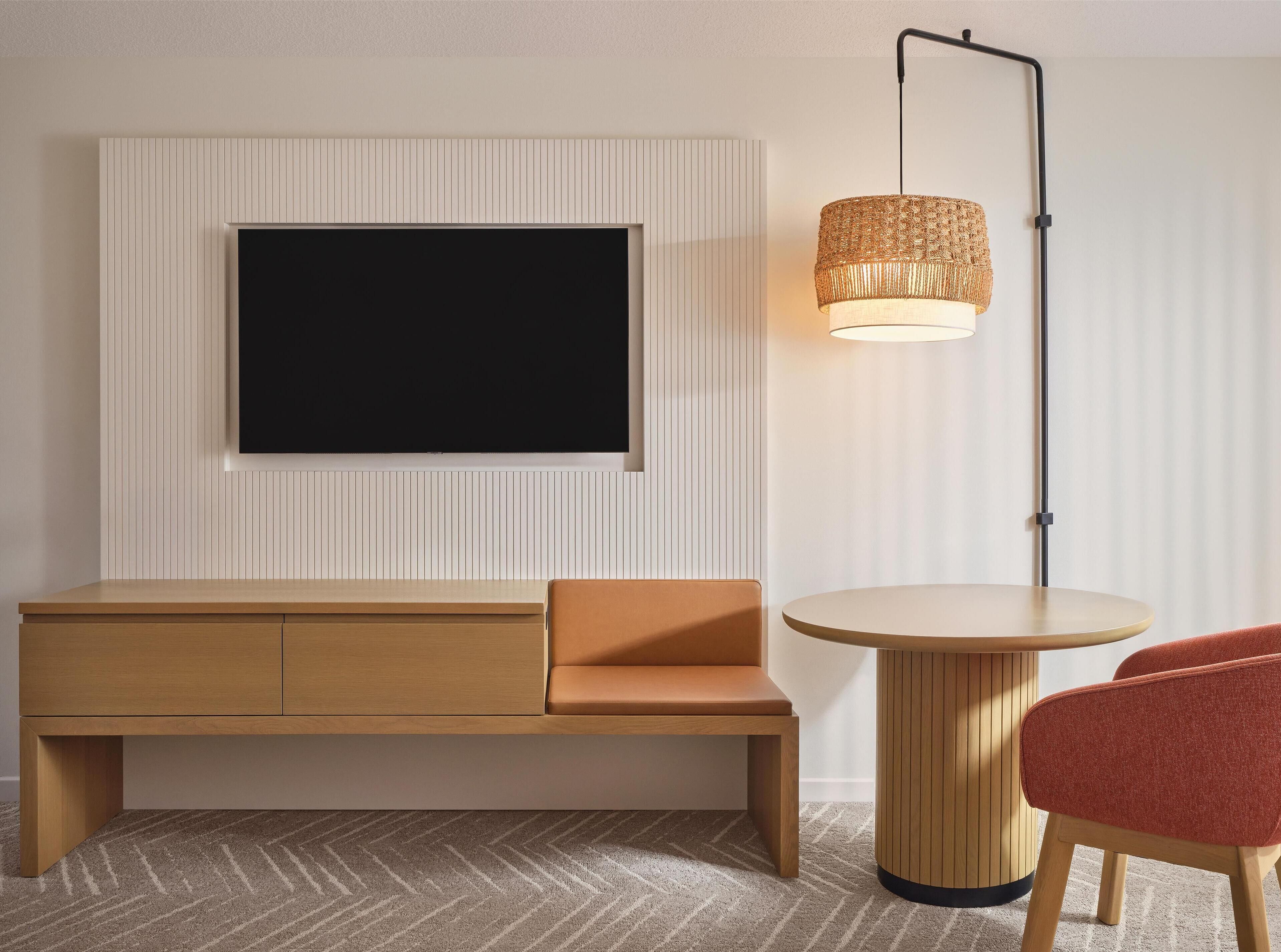Une chambre moderne avec un téléviseur à écran plat fixé à un mur blanc, un banc en bois doté d’un siège rembourré, une table ronde en bois et un lampadaire avec un abat-jour en tissu tressé.
