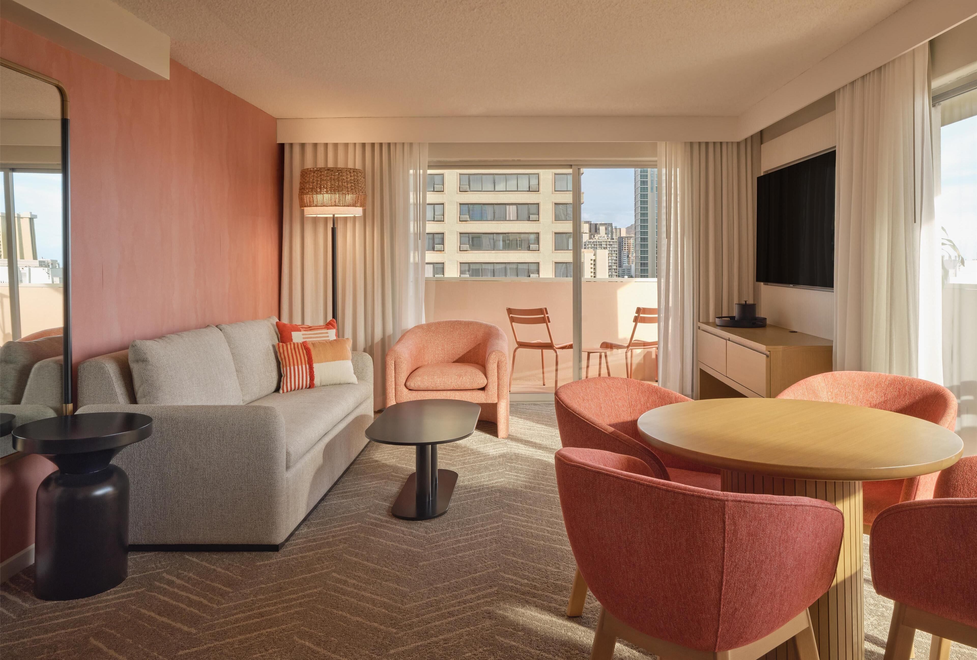 A modern hotel room with a sofa, armchairs, a dining table, a TV, and a balcony with a city view.