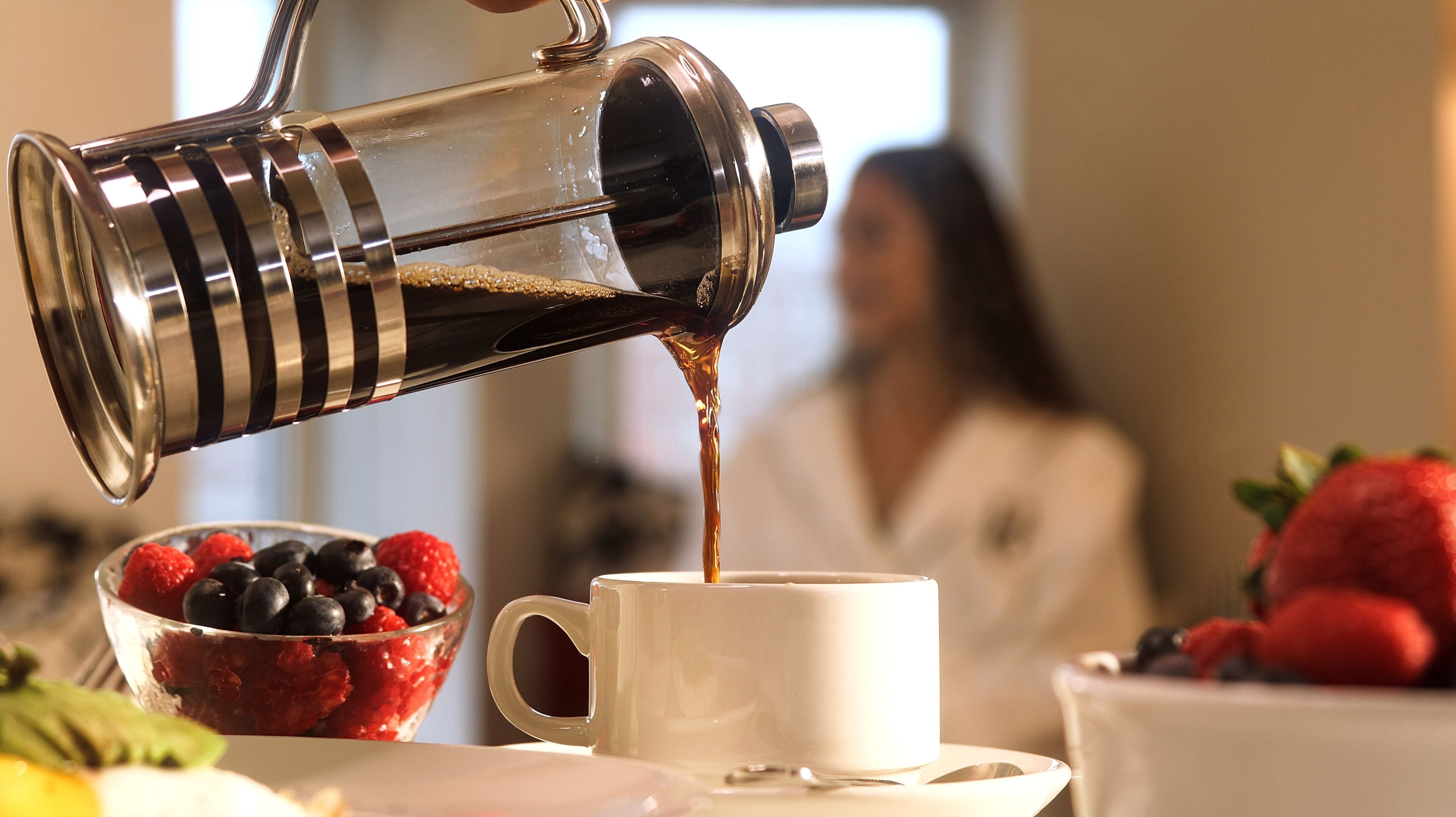 A coffee maker pouring coffee into a cup, a bowl of berries, and a person in the background.