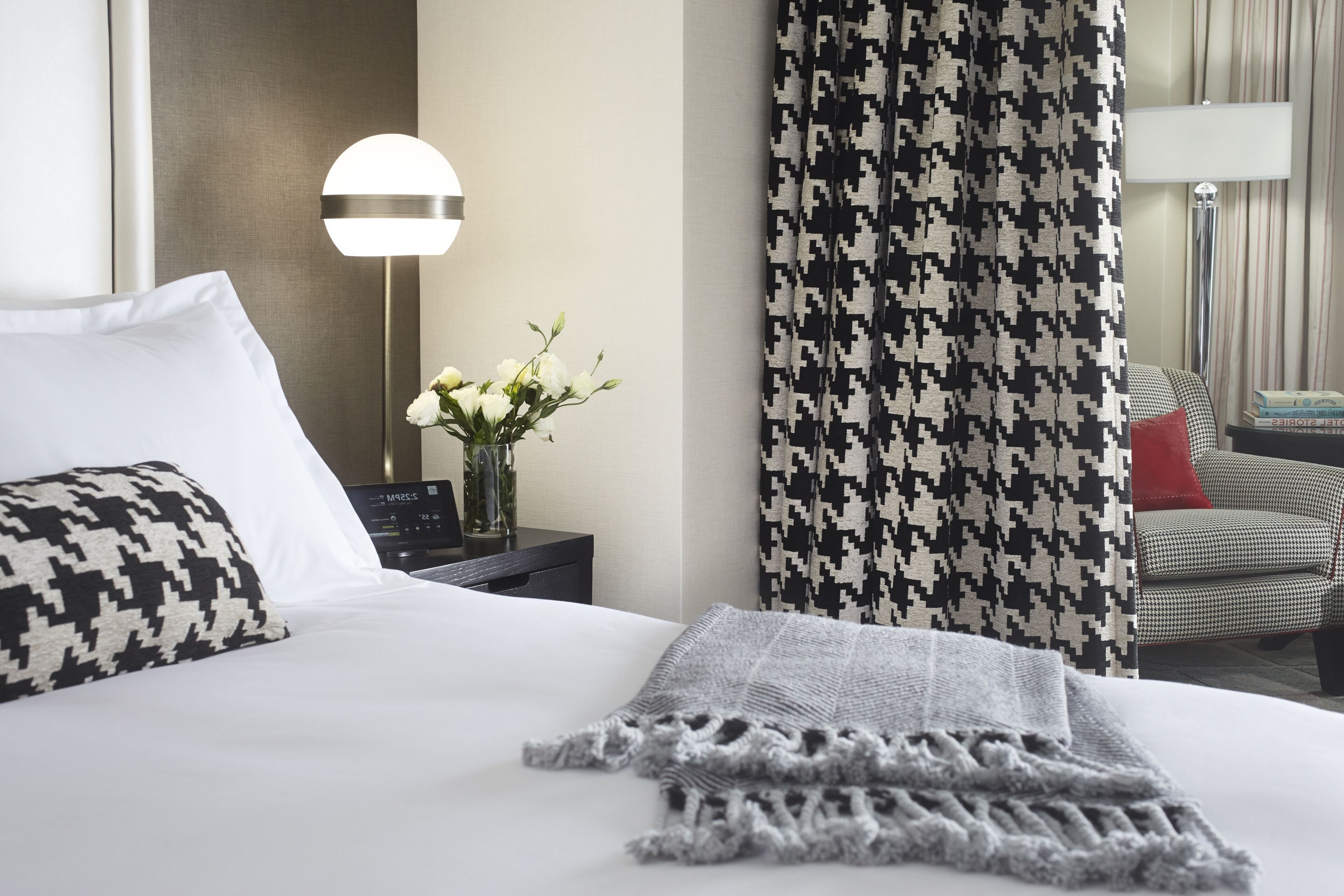 Una habitación de hotel con una cama bien hecha, una lámpara, una cortina de estampado pata de gallo y una silla con un cojín rojo.