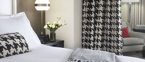 Una habitación de hotel con una cama bien hecha, una lámpara, una cortina de estampado pata de gallo y una silla con un cojín rojo.