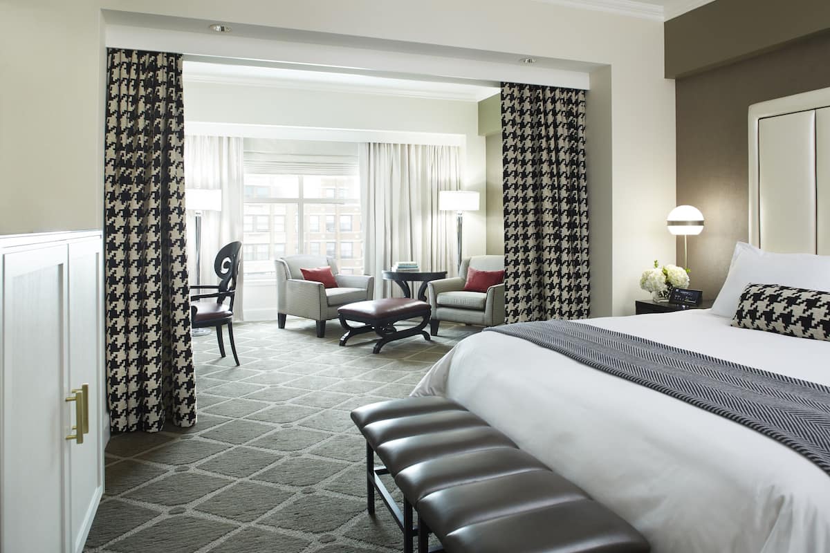 Habitación de hotel con una cama grande, un banco, una mesita pequeña y dos sillas. Tiene una ventana con cortinas, una lámpara y una mesita de noche con un jarrón de flores.