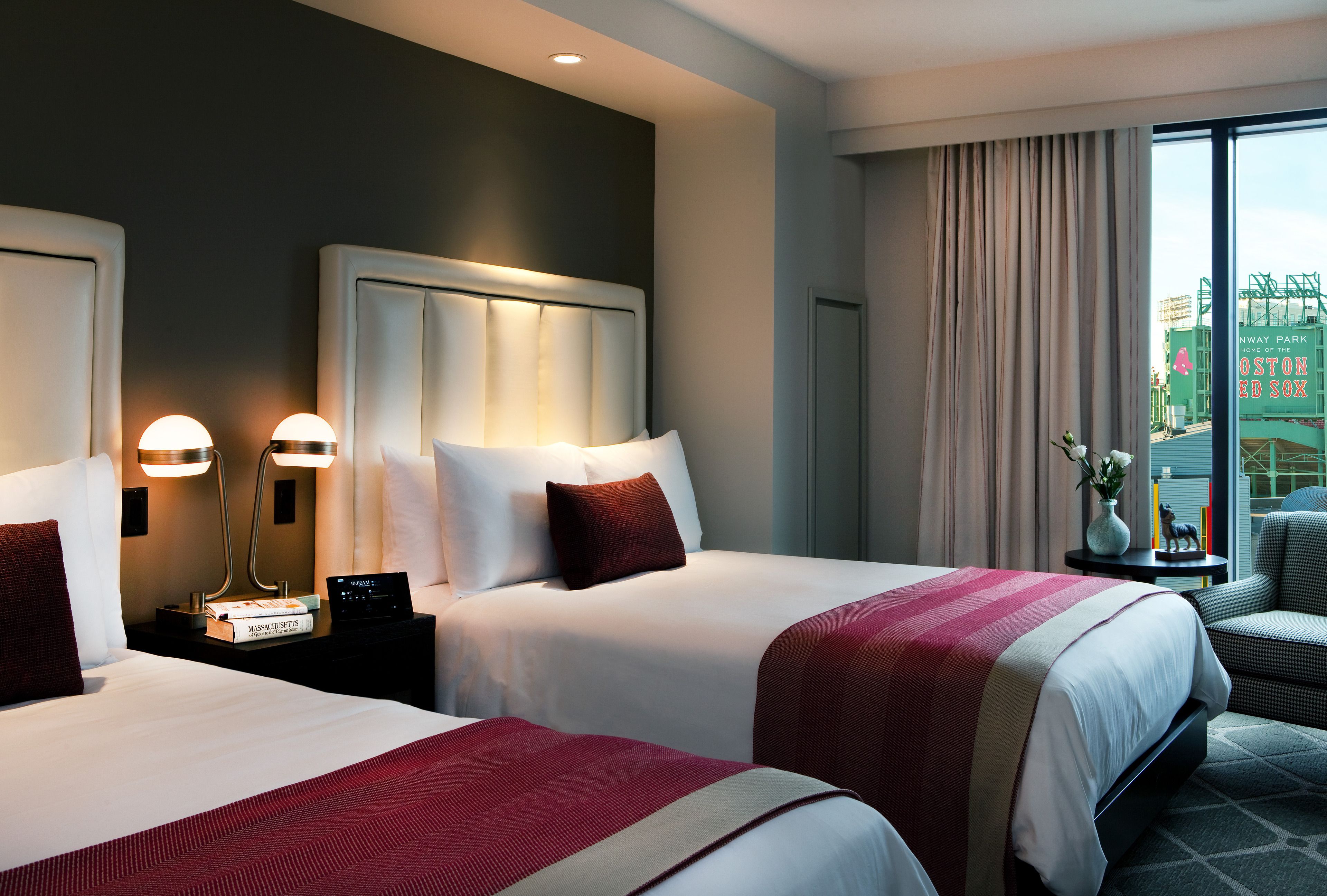 A hotel room with two beds, a nightstand with a lamp, a small table with a vase and a book, and a view of a building outside.