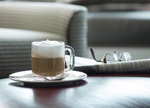 A clear glass cup of coffee on a saucer, a pair of glasses, and a newspaper on a table.