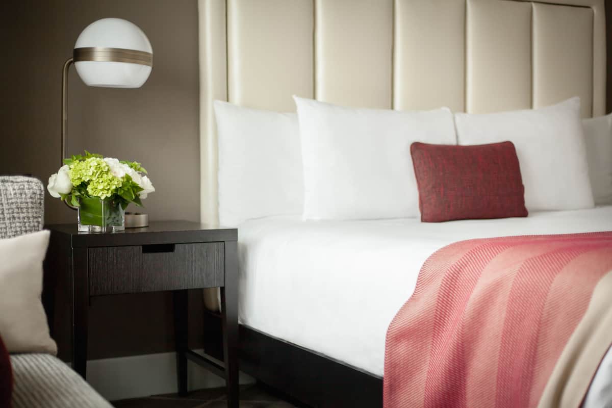 Una cama bien tendida con almohadas blancas y rojas, una lámpara de noche y un florero con flores sobre una mesita de noche oscura.