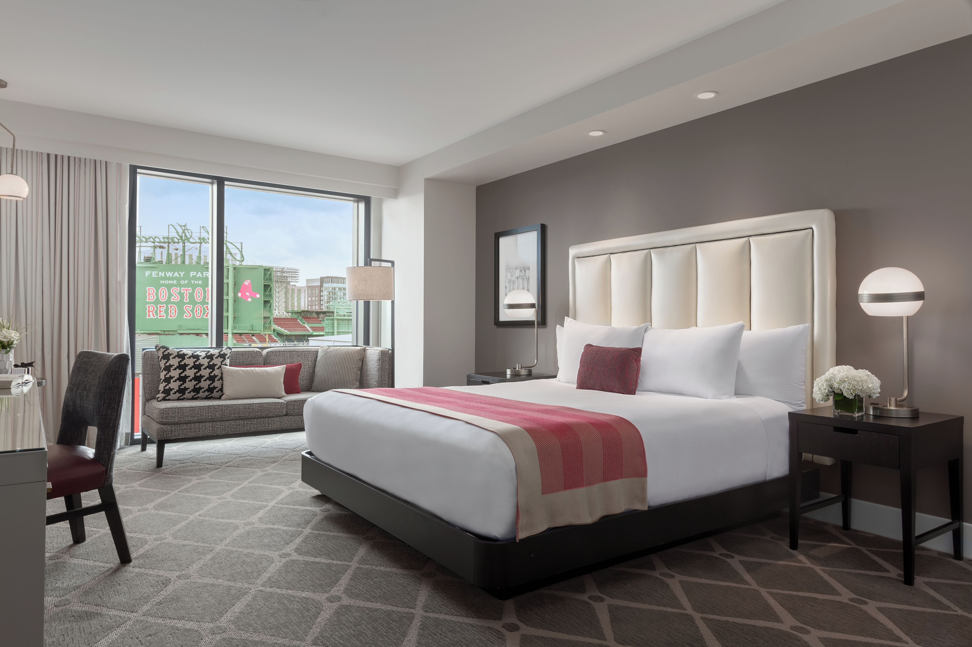 A modern hotel room with a large bed, a sofa, a desk, and a view of Fenway Park.