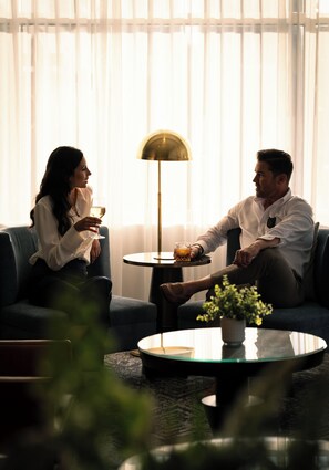 A man and a woman sitting on a blue sofa, holding glasses of a beverage. There is a small round table with a glass of orange juice and a potted plant in the foreground.