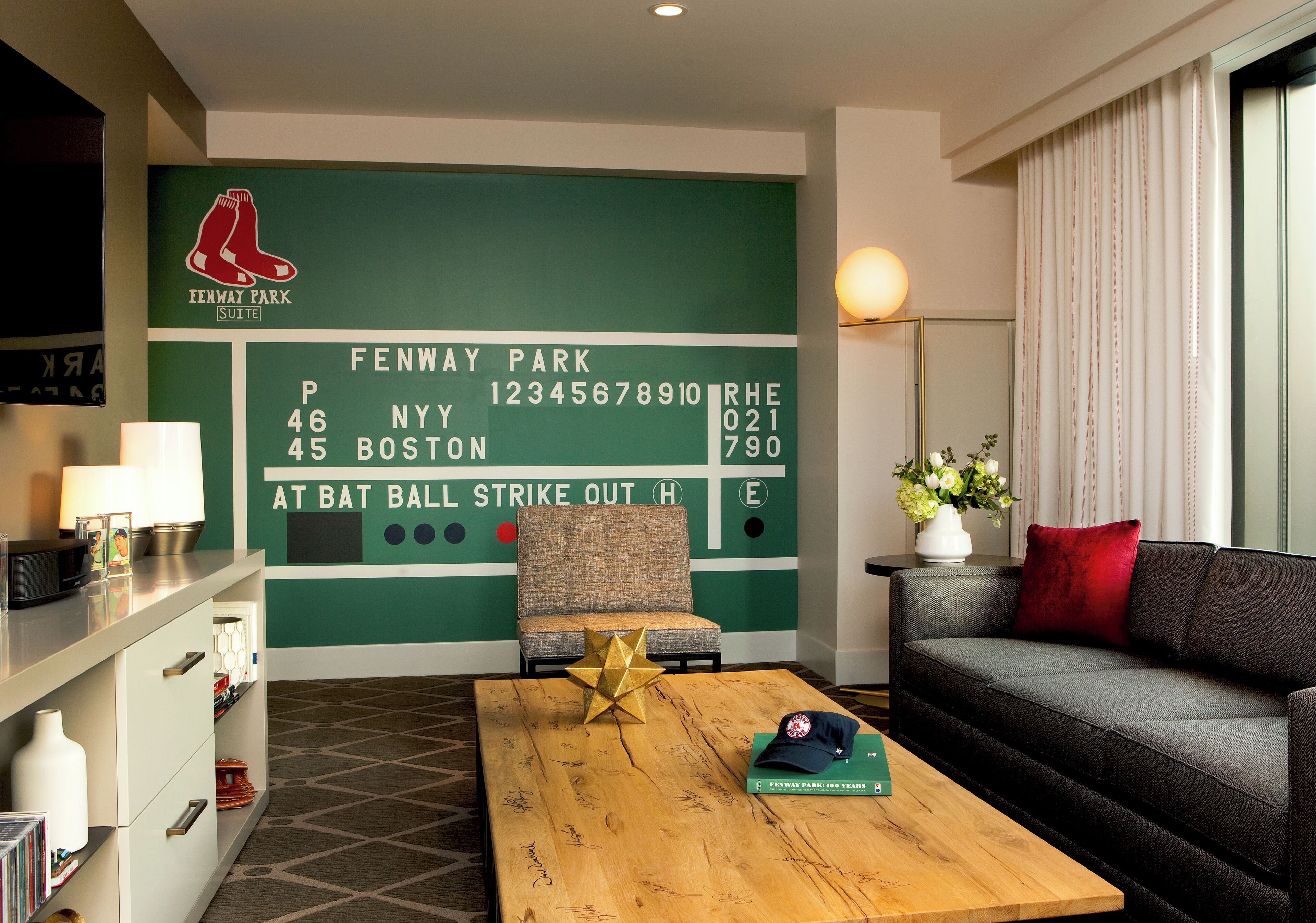 A hotel room with a green wall featuring a Boston Red Sox logo and a scoreboard. There is a sofa, a coffee table with a baseball cap and a book, and a lamp.