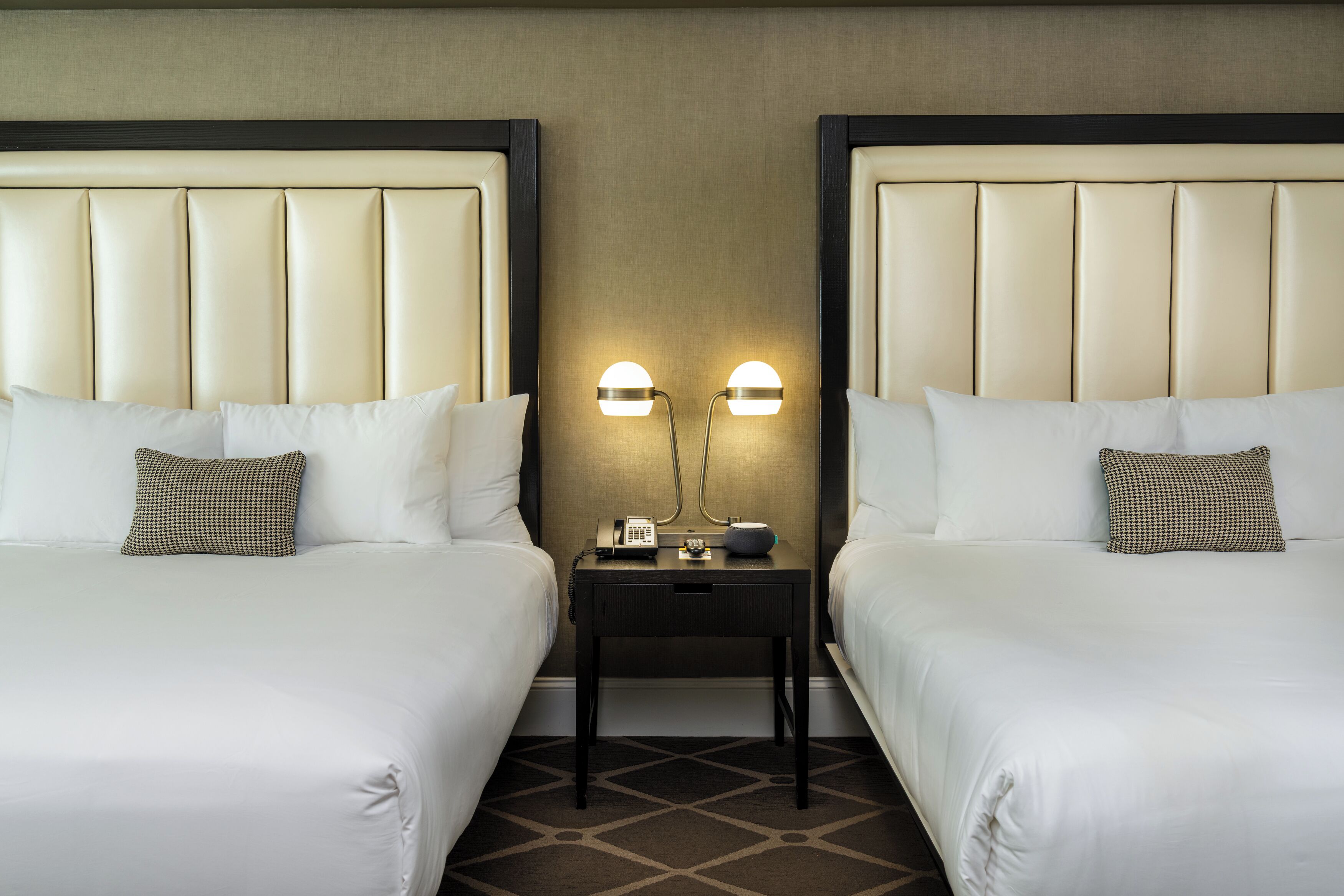 A hotel room with two beds, a nightstand with a lamp, and a wall-mounted phone.