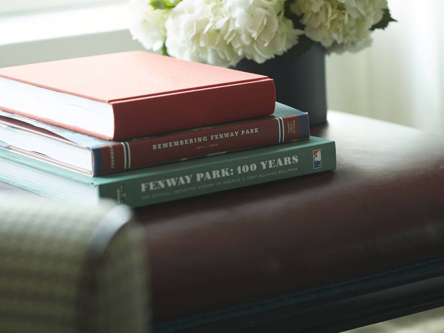 A stack of books on a leather couch with a vase of white flowers in the background.