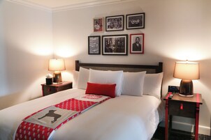 Uma cama bem arrumada com um cobertor vermelho e branco, duas luminárias de mesa e uma série de fotografias emolduradas na parede.