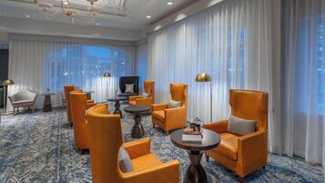 Un vestíbulo de hotel con una araña, ventanas amplias con cortinas y sillones tapizados en color naranja.