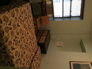 A room with a floral-patterned bedspread, a small table with a chair, a window with blinds, and a framed picture on the wall. - Park Savoy (New York)