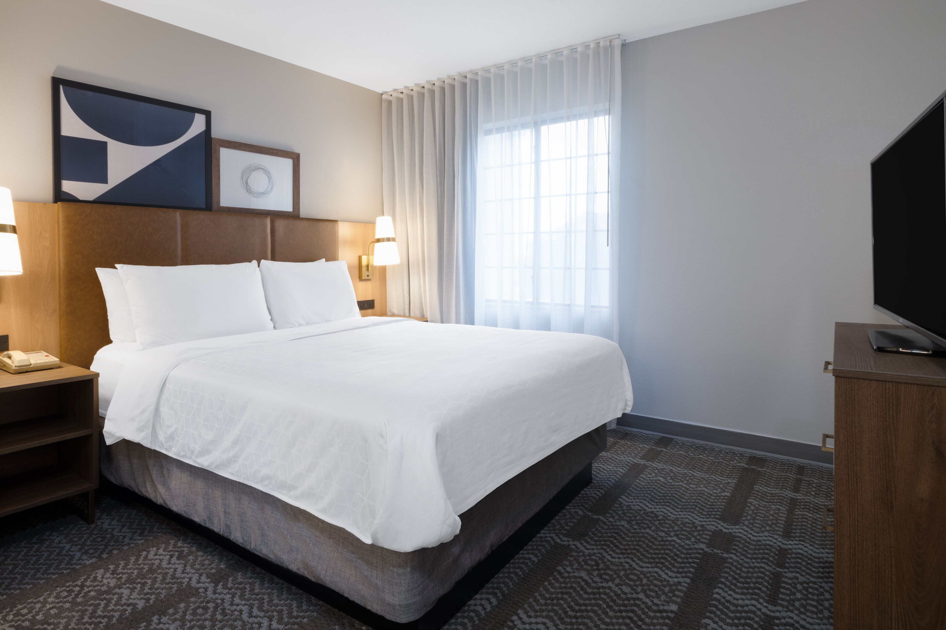A hotel room with a large bed, a flat-screen TV, a nightstand with a lamp, and a window with sheer curtains.