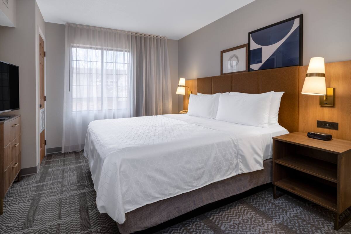 A hotel room with a large bed, two bedside lamps, a TV, and a window with sheer curtains.