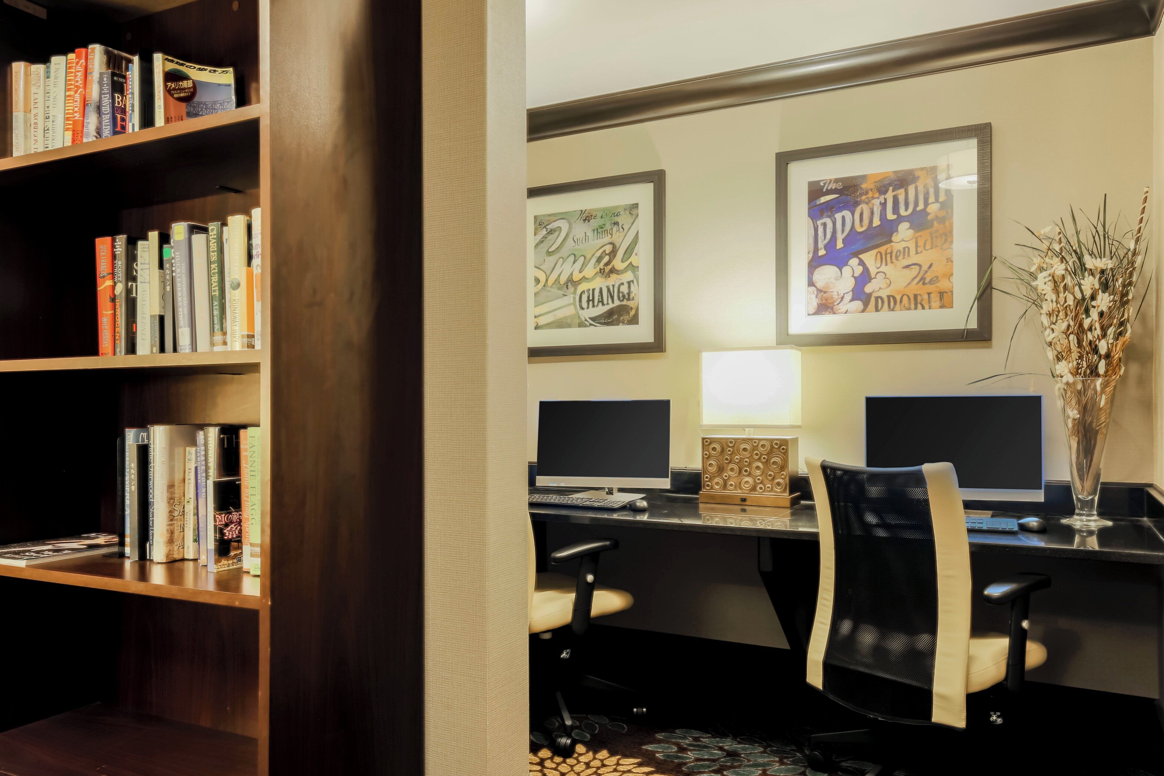 A room with a desk, two computer monitors, a lamp, a chair, and a bookshelf filled with books.