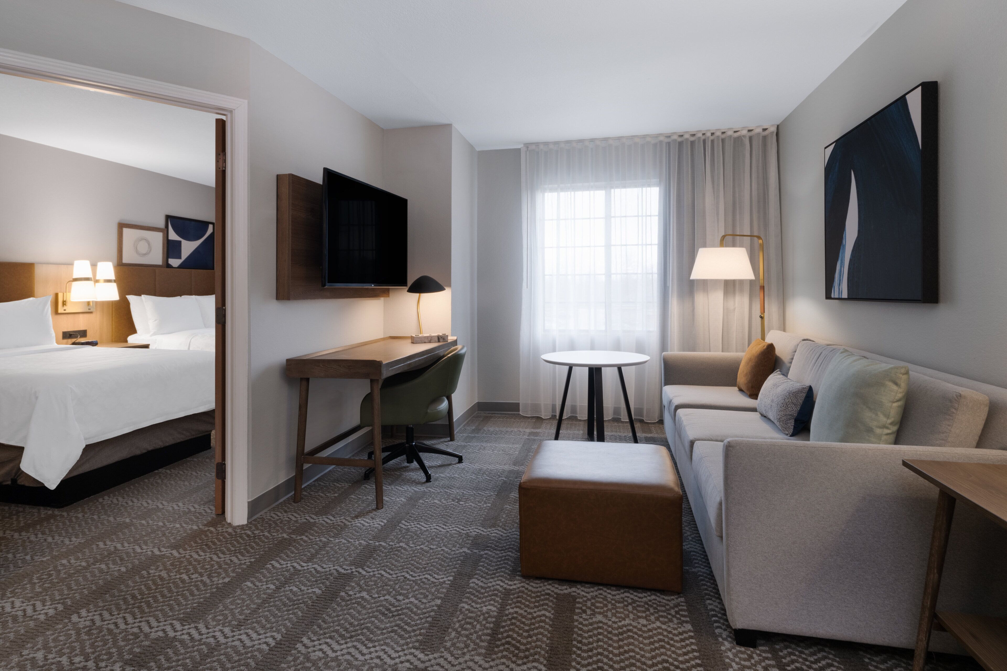 A hotel room with a bed, desk, chair, sofa, ottoman, and a TV mounted on the wall.