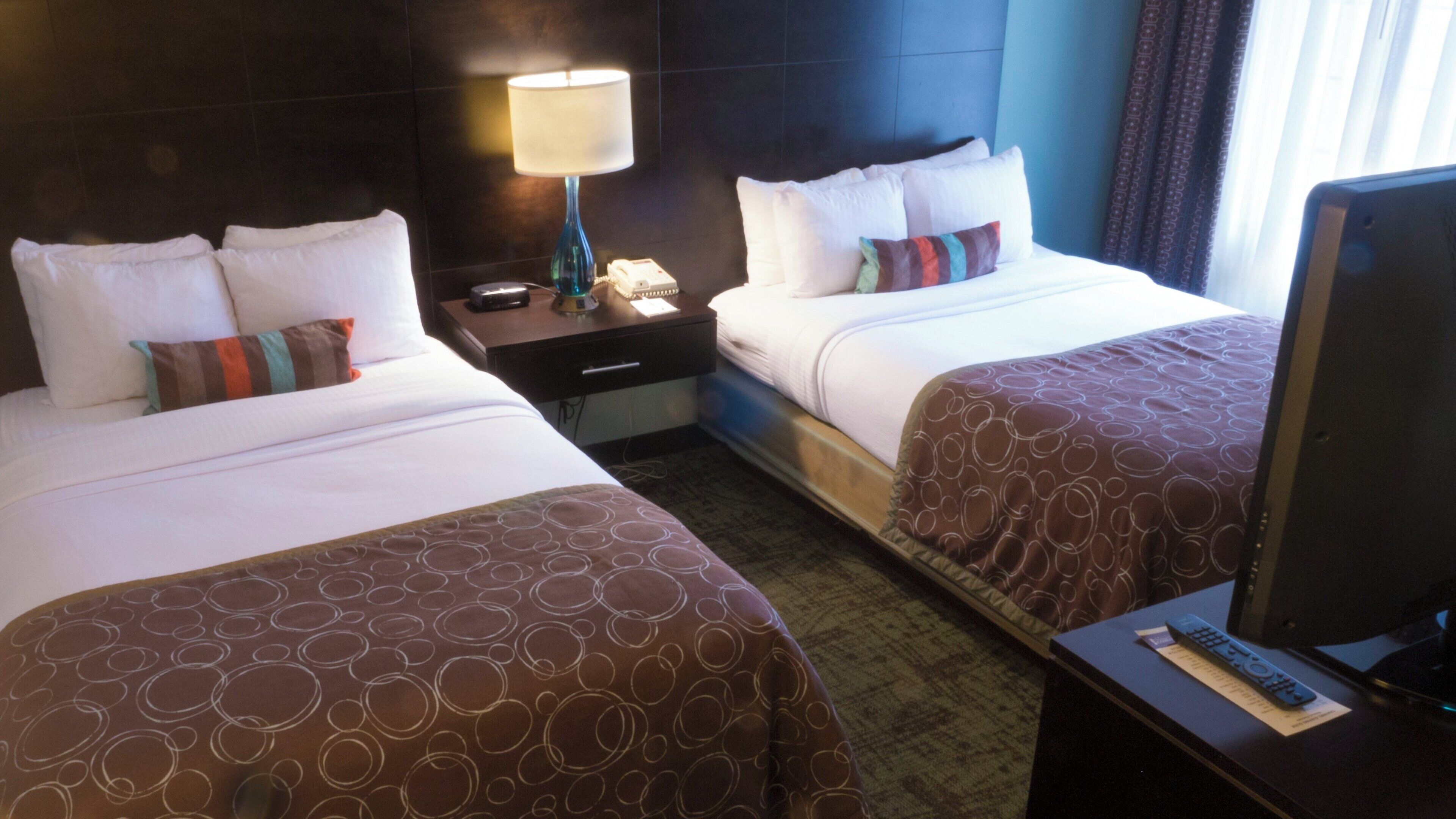 A hotel room with two beds, a nightstand, a lamp, a television, and a remote control.