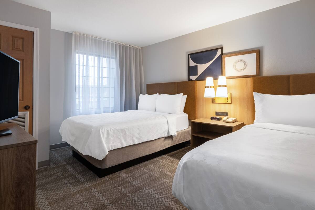 A hotel room with two beds, a TV, a desk, a lamp, and a window with curtains.