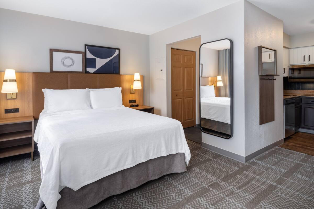 A hotel room with a large bed, bedside lamps, a mirror, a wooden headboard, a framed picture, and a kitchen area visible through an open door.