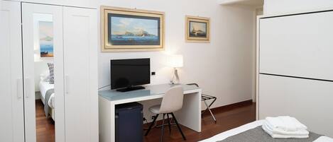 Een hotelkamer met een bed, een bureau met een computer, een stoel, een lamp en twee afbeeldingen in lijsten aan de muur.