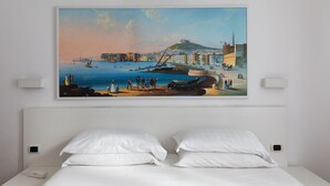 Une chambre d’hôtel avec un lit, des oreillers blancs et un tableau représentant un paysage urbain côtier accroché au mur.
