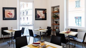 Un espace de restauration moderne avec des tables blanches et des chaises noires, des œuvres d’art encadrées aux murs et une vue sur les bâtiments à l’extérieur.