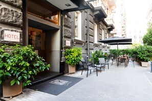 An outdoor hotel entrance with a doormat, potted plants, and outdoor seating. - Rex Lifestyle Hotel (Naples)