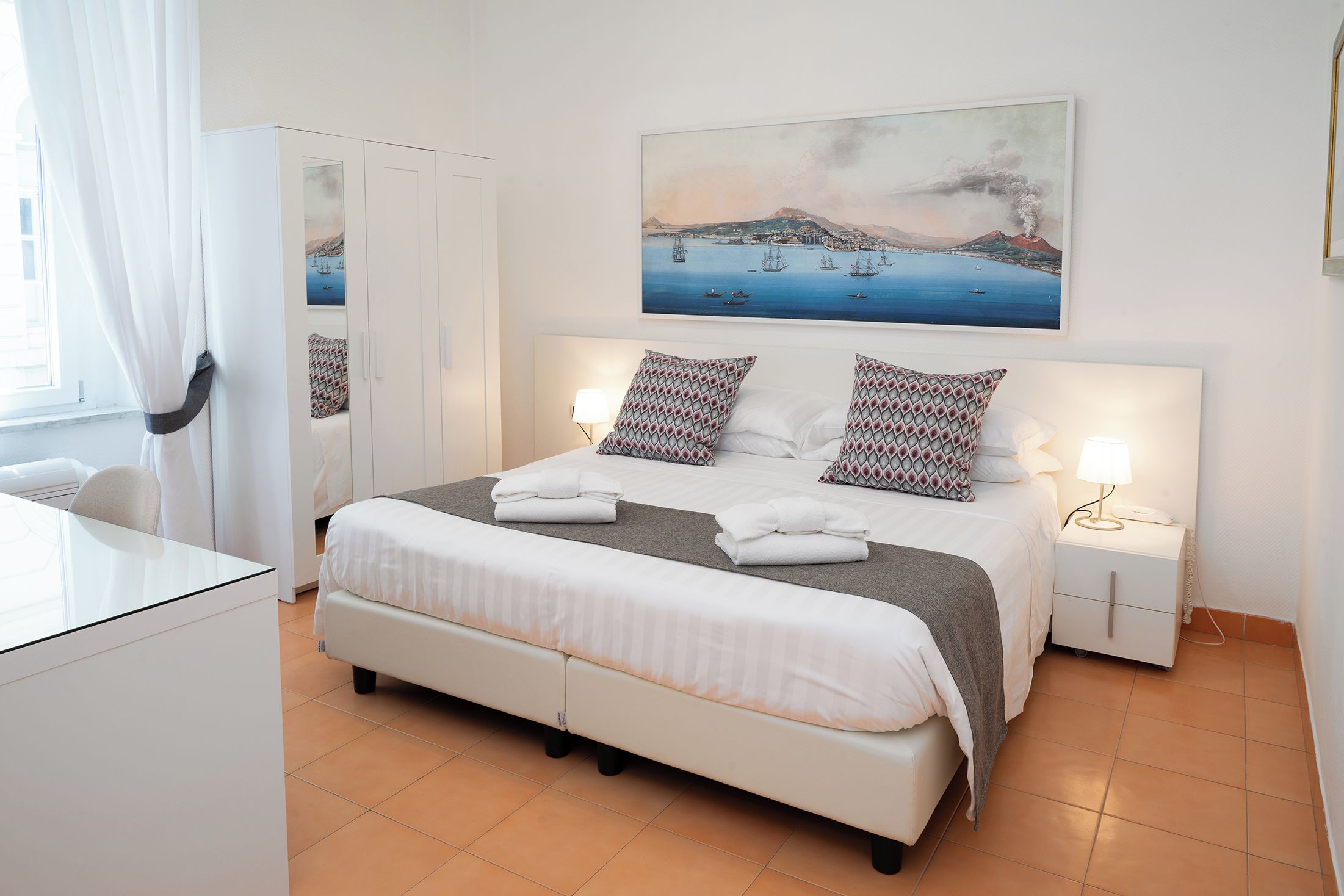 A hotel room with a bed, bedside tables, a desk, and a painting of a coastal scene.
