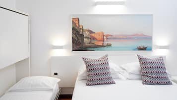 A hotel room with a bed, two patterned pillows, a painting of a coastal scene, and a wall-mounted light fixture.