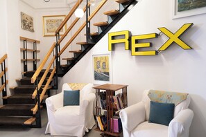 A staircase with wooden steps and black railings, a white armchair with a blue pillow, another white armchair with a floral pillow, and a bookshelf with books. - Rex Lifestyle Hotel (Naples)