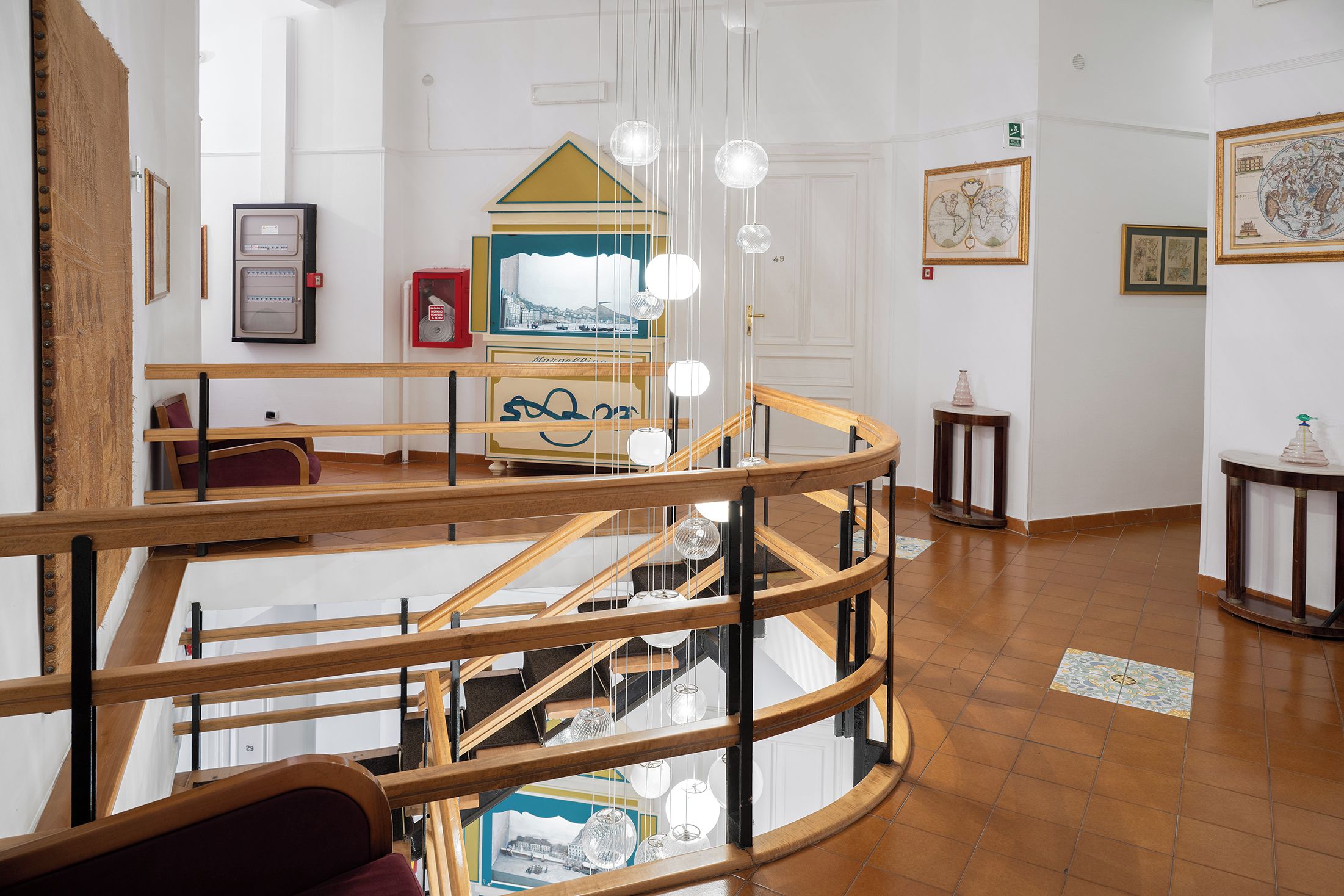 Ein mehrgeschossiges Interieur mit einer Holztreppe, hängenden Pendelleuchten und gerahmten Kunstwerken an den Wänden.