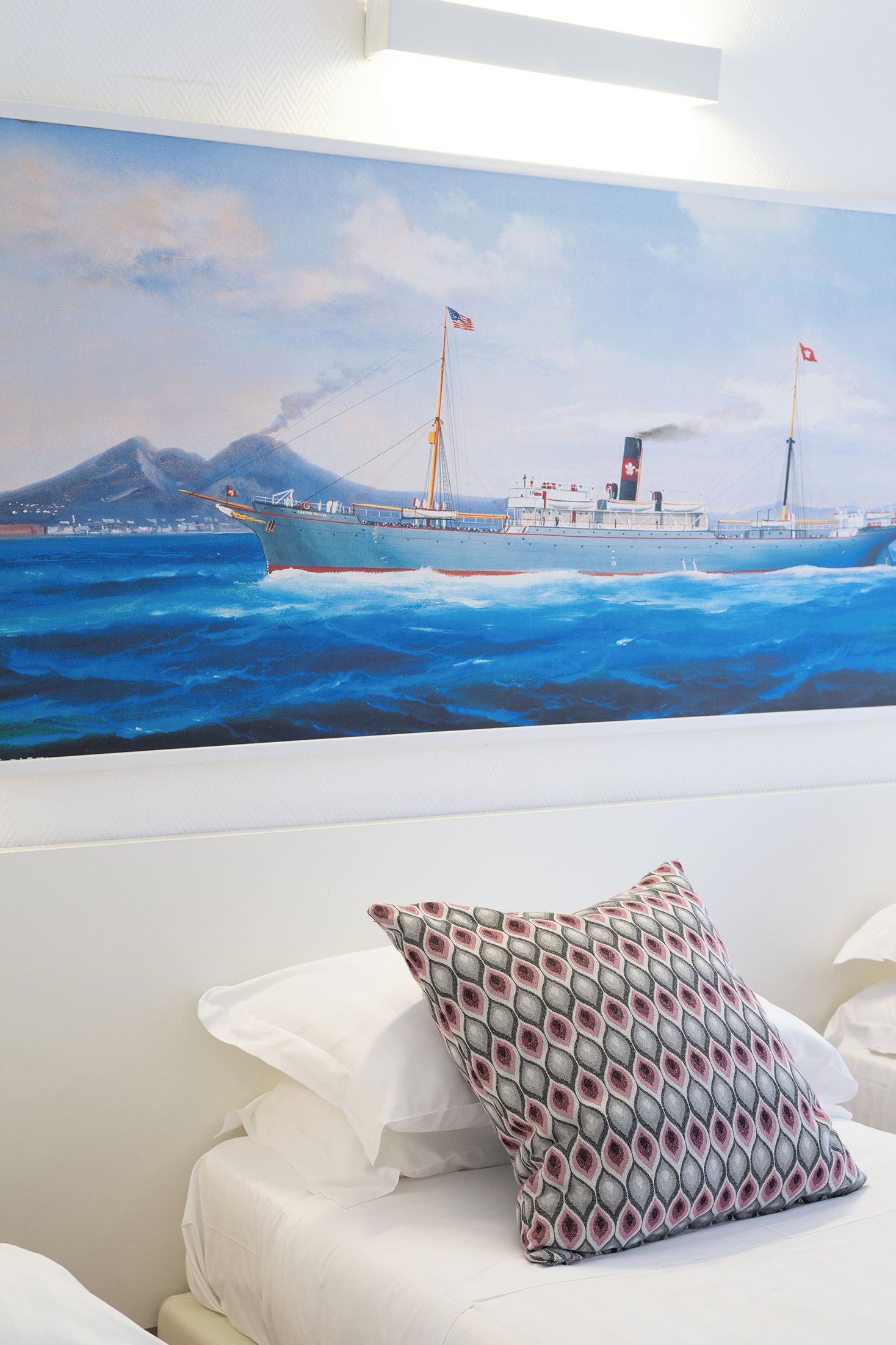 A room with a large painting of a ship on the ocean, a white bed with a patterned pillow, and a white wall.
