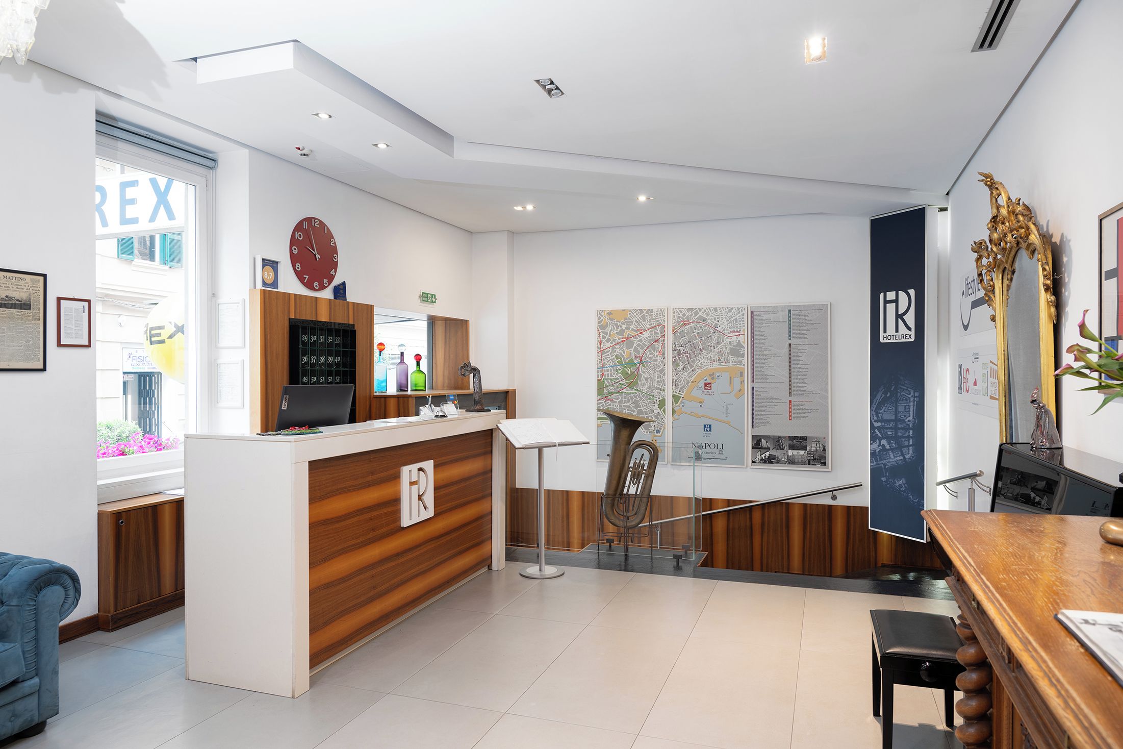 A hotel reception area with a wooden reception desk, a clock, a map, and a large window with a view of the outside.