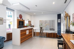 A hotel reception area with a wooden reception desk, a clock, a map, and a large window with a view of the outside. - Rex Lifestyle Hotel (Naples)