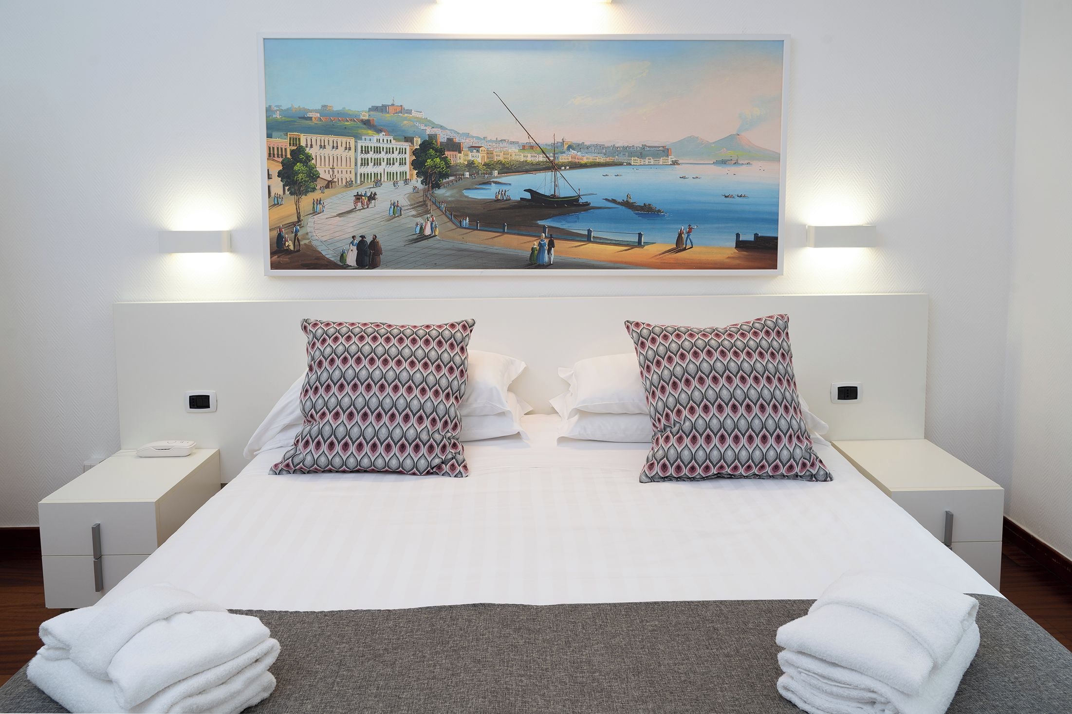 A hotel room with a bed, two patterned pillows, a nightstand, a bedside table, a wall-mounted painting of a coastal scene, and a neatly folded towel on the bed.